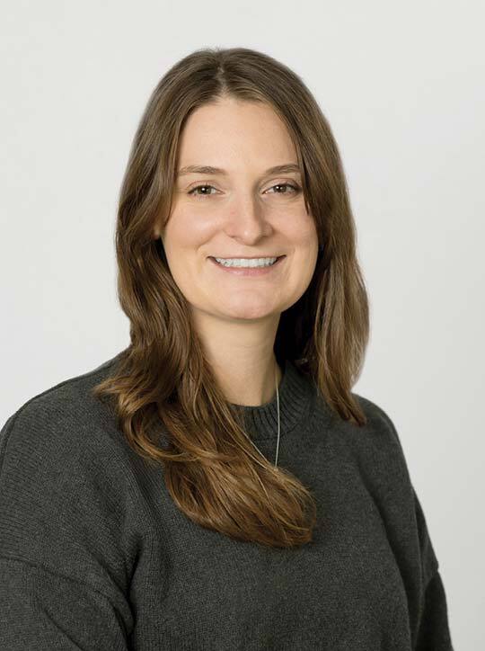 Smiling woman with long brown hair wearing a dark sweater in a professional headshot against a light background.