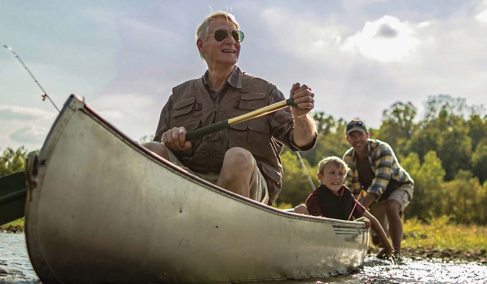Older man paddling a canoe with a young child in front and another adult behind, enjoying a sunny day outdoors.