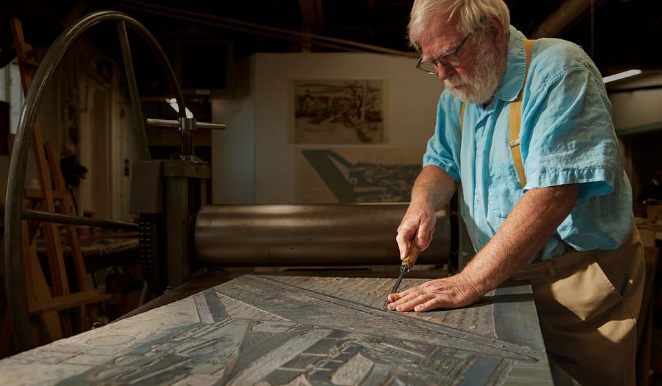 Older man carving a woodblock at a printmaking press inside an art studio.