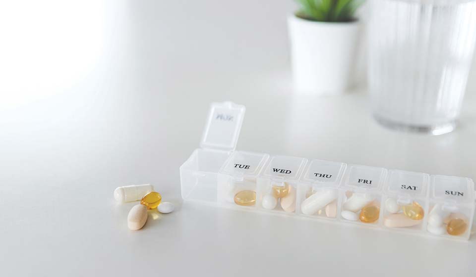 Clear weekly pill organizer filled with vitamins and supplements on a white table.