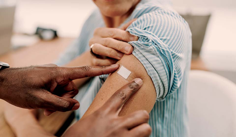 Close-up of healthcare worker pointing to a small bandage on a patient’s upper arm after vaccination.