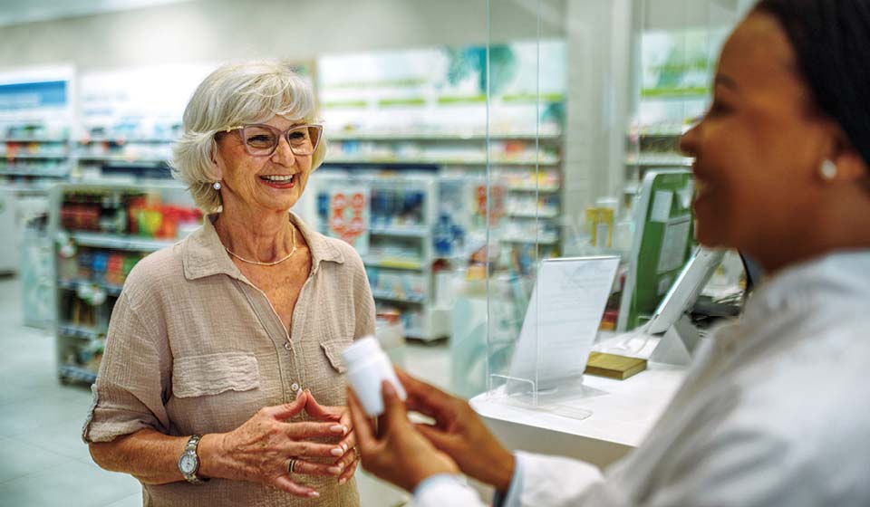 Pharmacist handing a prescription bottle to a smiling older woman at a pharmacy counter.