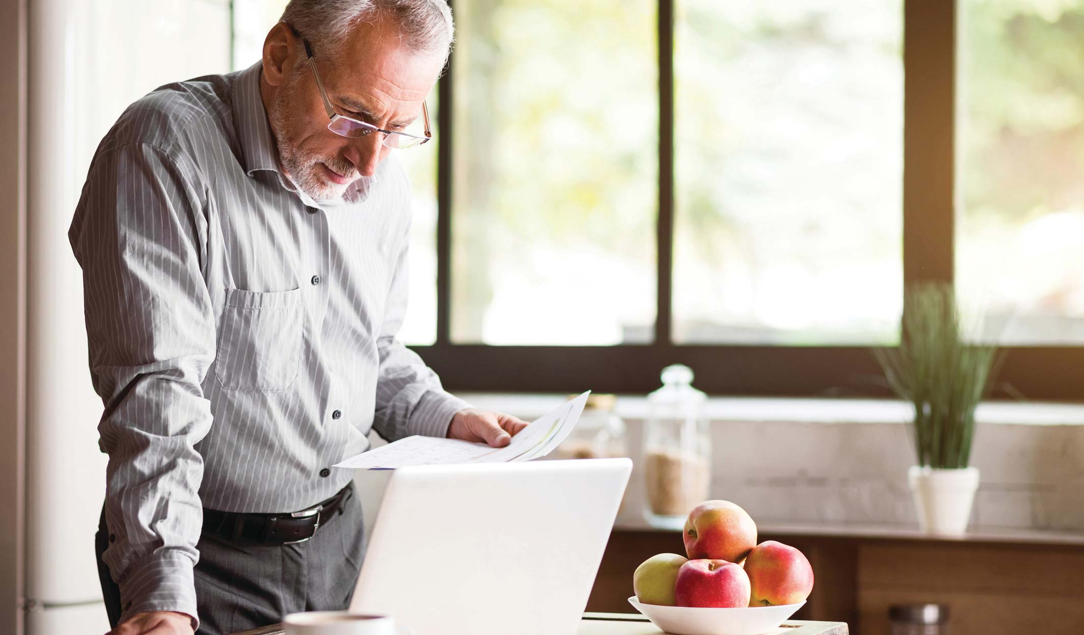 Senior man standing at a kitchen counter using a laptop.