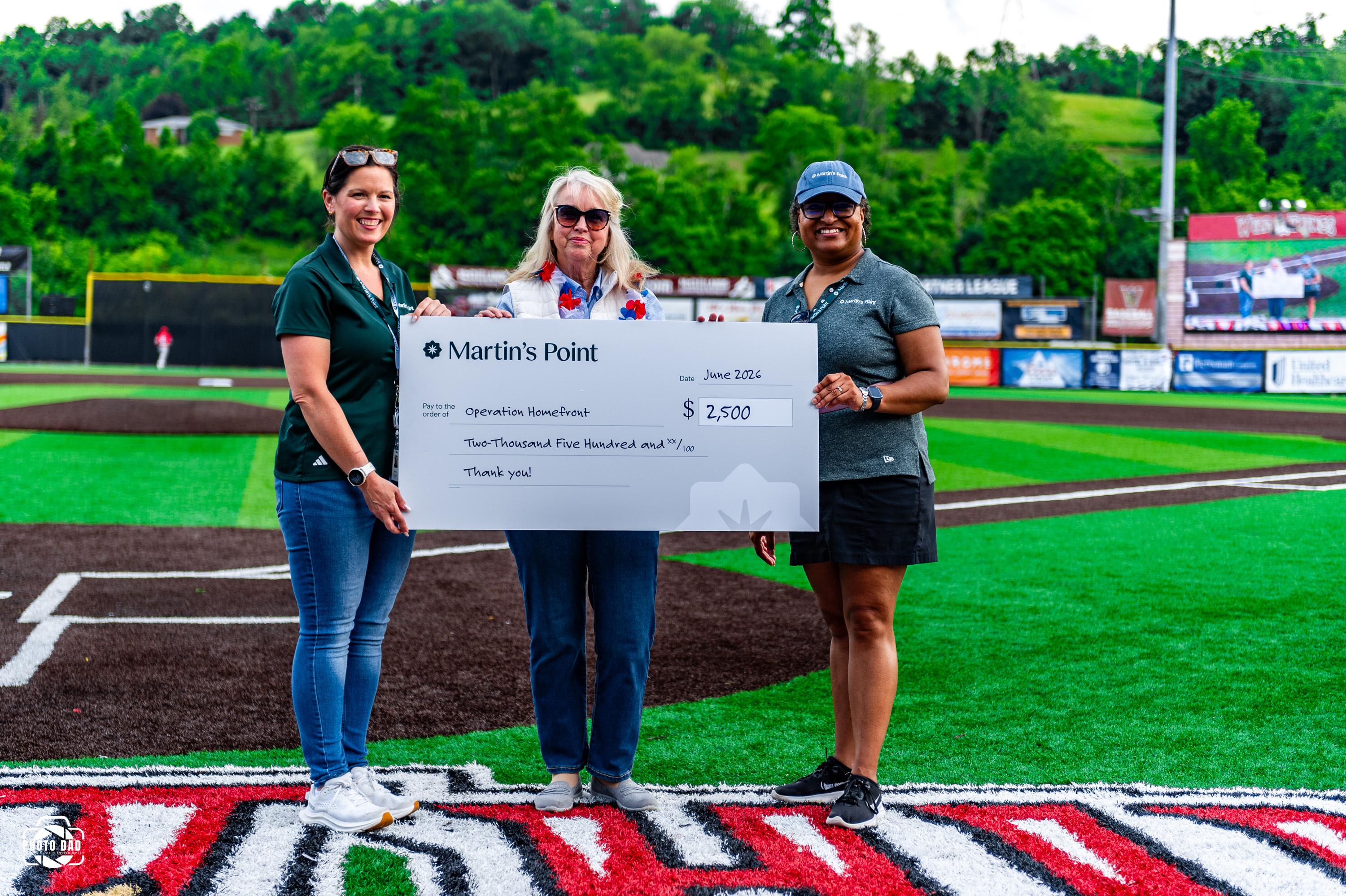 Three individuals hold a Martin’s Point donation check for Operation Homefront on a baseball field during a military appreciation event.