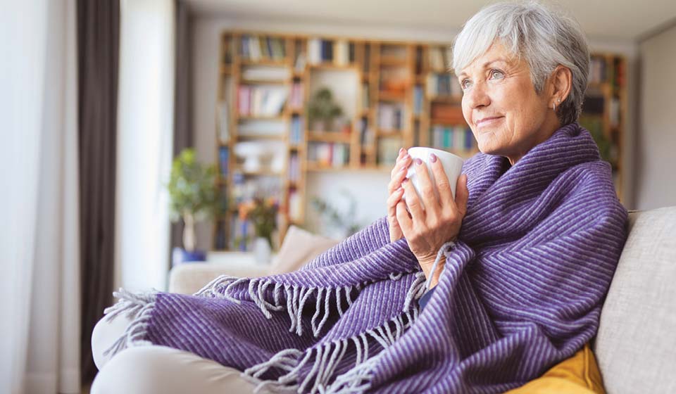 Older woman wrapped in a purple blanket holding a warm drink while relaxing at home.