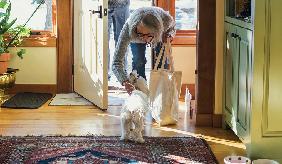 Older woman entering her home and greeting a small white dog with a grocery bag in hand.