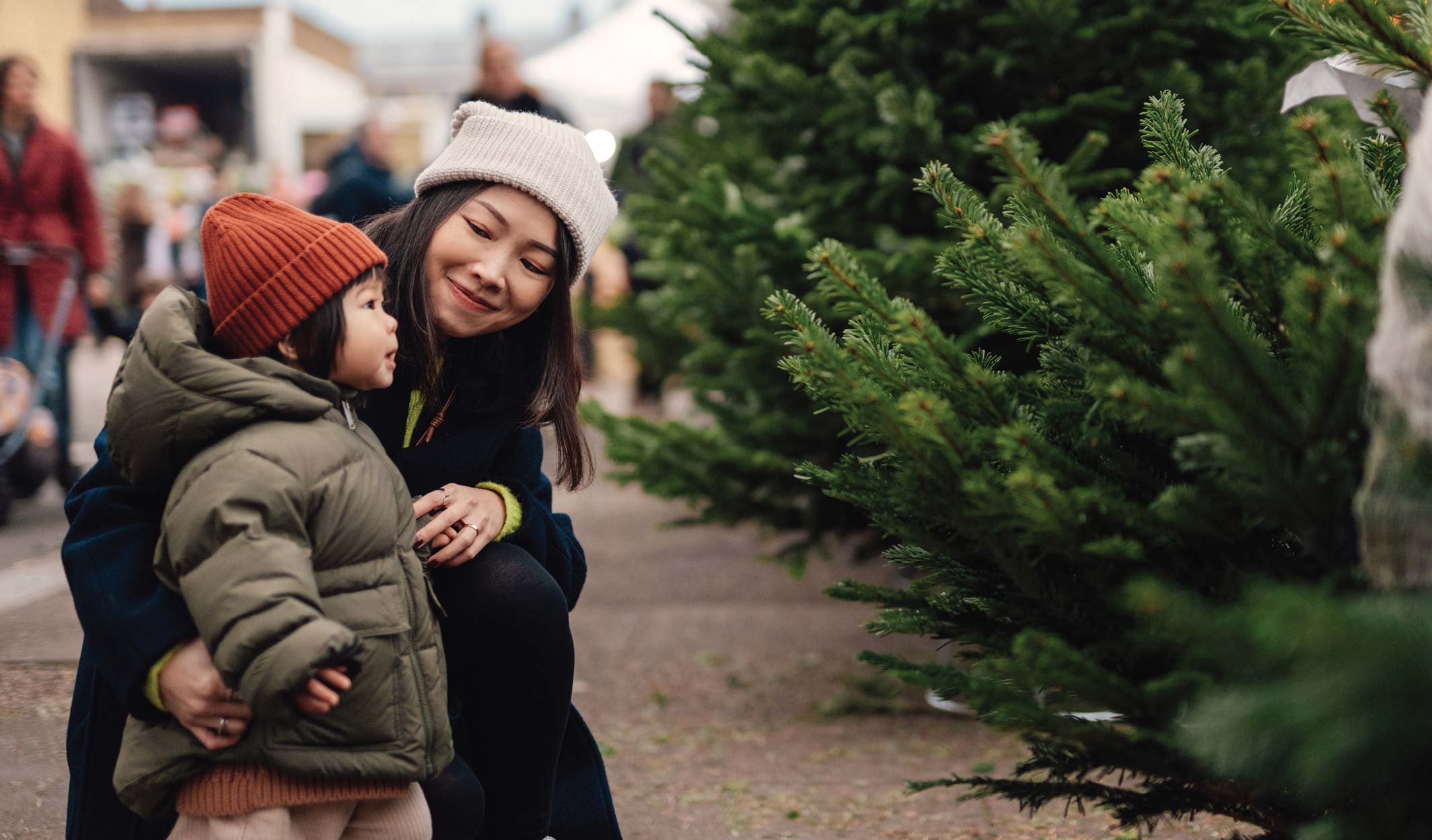 Mother holding her child while looking at Christmas trees outdoors.