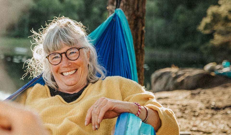 Smiling older woman relaxing in a blue hammock near a lake on a sunny day.