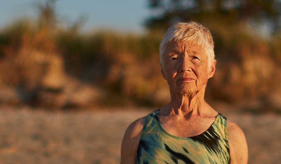 Elderly woman standing on a beach in a swimsuit at sunset, looking thoughtfully at the camera.
