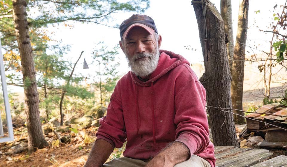 Older man in a red hoodie sitting outside in a wooded area, smiling warmly.