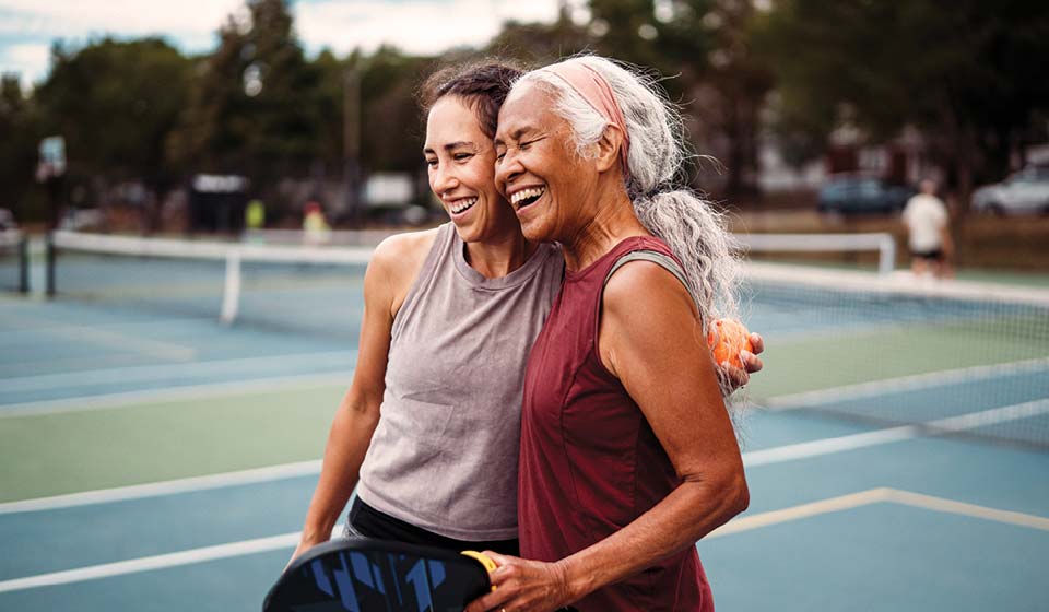 Two women smiling and hugging on a tennis court after playing a match.