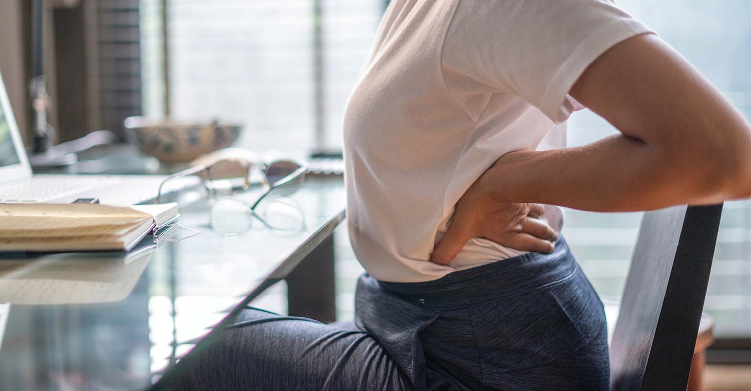 Person holding their lower back in pain while sitting at a table with a laptop and notebook.
