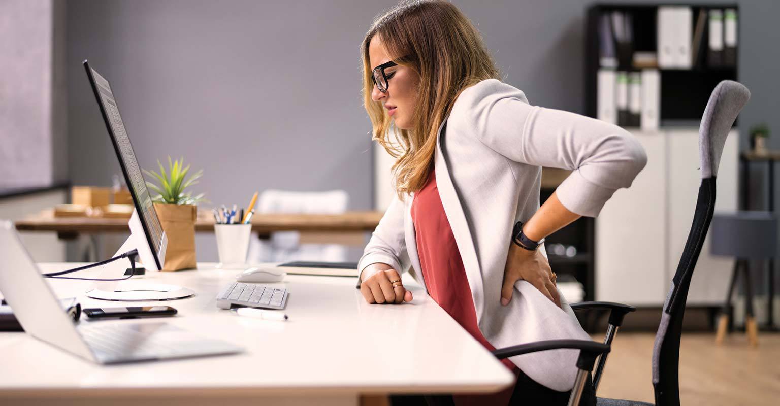 Woman sitting at her desk holding her lower back in discomfort while working on a computer.