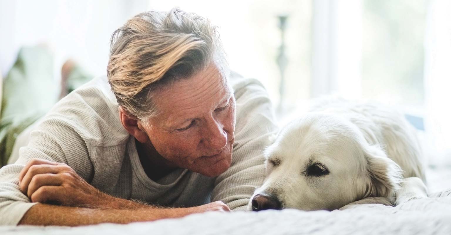 Older man lying on a bed beside his dog, showing quiet companionship.