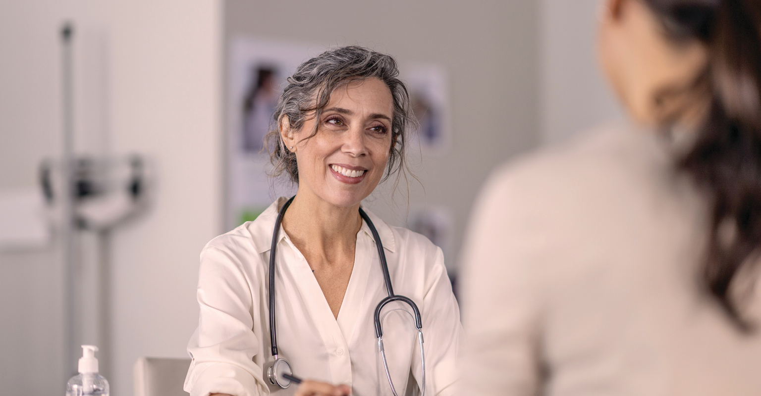 Nurse sitting at a desk, smiling while speaking with a patient.