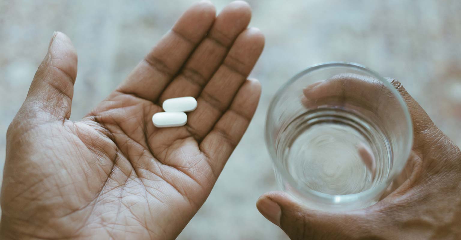 Person holding two pills in one hand and a glass of water in the other.