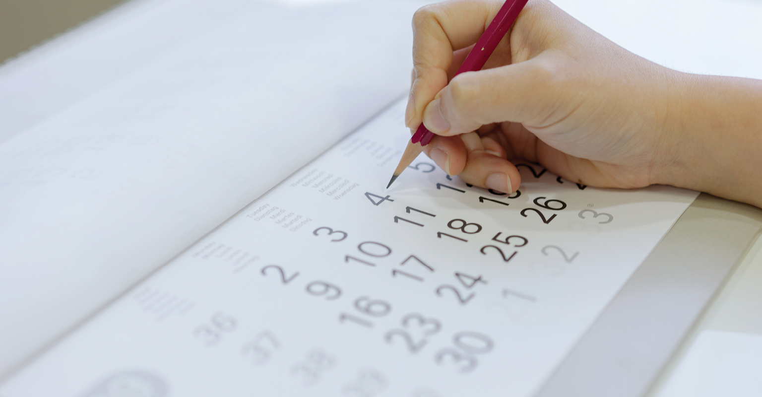 Hand marking a date on a paper calendar with a pencil.