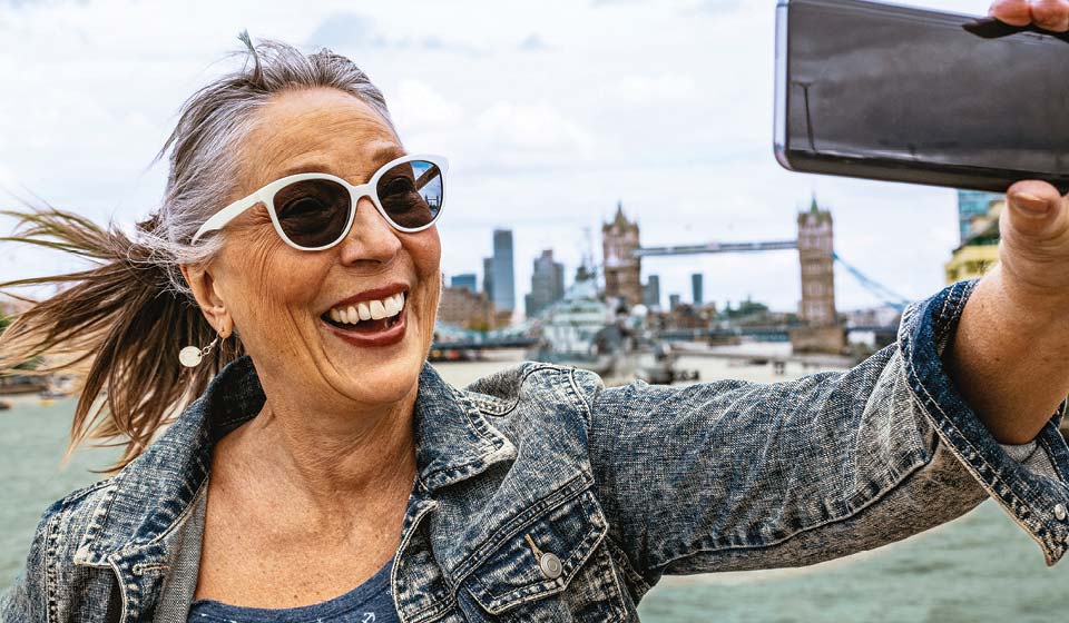 Smiling woman in sunglasses taking a selfie in front of the Tower Bridge in London.