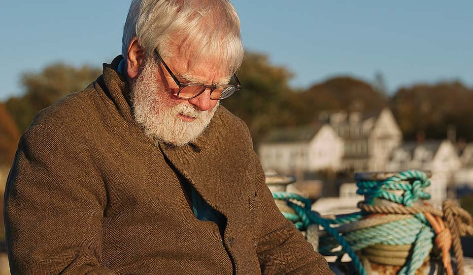Older man with white hair and beard sitting near a dock surrounded by colorful ropes and coastal buildings.