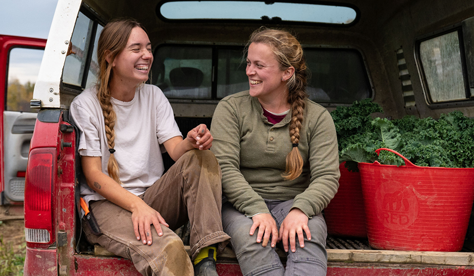 Two women farmers laughing together while sitting in a truck bed with fresh kale.